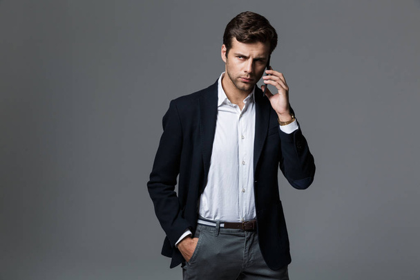 Photo of a handsome young concentrated business man isolated over grey wall background talking by mobile phone. - Φωτογραφία, εικόνα
