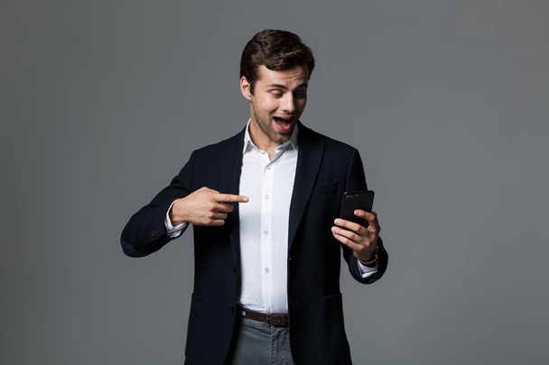 Portrait of a happy young businessman dressed in suit isolated over gray background, pointing finger at mobile phone - Foto, Bild