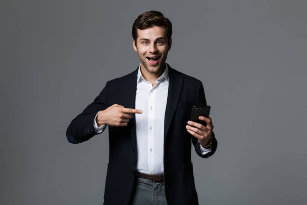 Πορτρέτο του ένα χαρούμενο νεαρός επιχειρηματίας ντυμένος με κοστούμι που απομονώνονται σε γκρίζο φόντο, που δείχνει δάχτυλο σε κινητό τηλέφωνο - Φωτογραφία, εικόνα