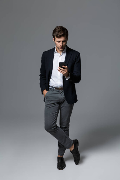 Full length portrait of a confident young businessman dressed in suit isolated over gray background, standing, holding mobile phone - Foto, imagen