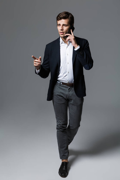 Full length portrait of a handsome young businessman dressed in suit isolated over gray background, walking, talking on mobile phone, pointing - Photo, image