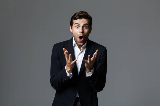 Portrait of an excited young businessman dressed in suit isolated over gray background, celebrating success - 写真・画像
