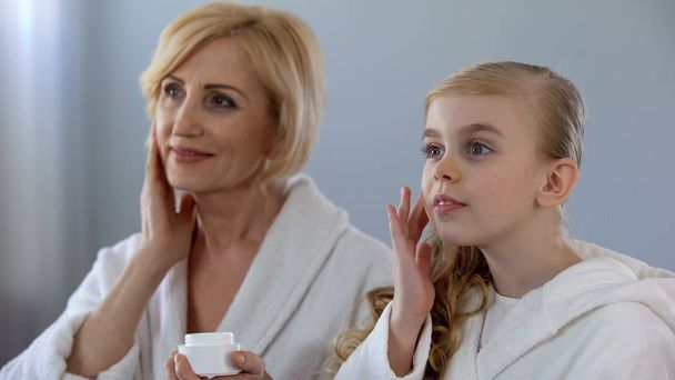 Pretty grandmother and granddaughter applying face cream and smiling, skin care - Фото, изображение