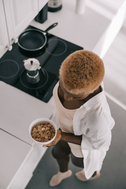 overhead view of african american woman holding bowl with cornflakes in white kitchen  - Foto, afbeelding