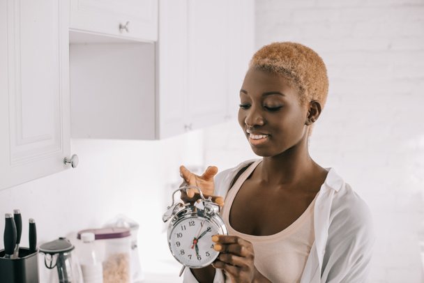 beautiful african american woman with short hair holding clock in white citchen  - Фото, зображення
