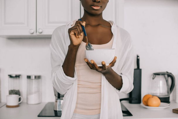 cropped view of african american woman holding spoon and bowl with cornflakes in white kitchen  - Foto, imagen