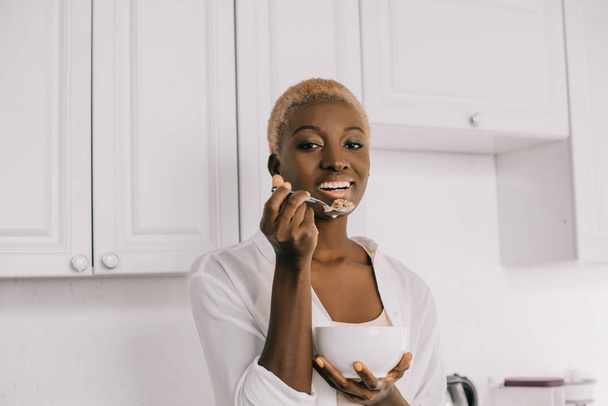 cheerful african american woman eating cornflakes in white kitchen  - Valokuva, kuva