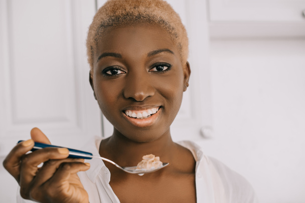 Nahaufnahme einer fröhlichen afrikanisch-amerikanischen Frau, die einen Löffel mit Cornflakes in der weißen Küche hält  - Foto, Bild