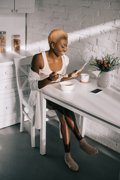 cheerful african american woman reading newspaper and having breakfast in white kitchen  - Fotó, kép