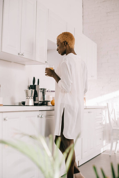 african american woman with short hair holding glass of orange juice in white kitchen  - Φωτογραφία, εικόνα