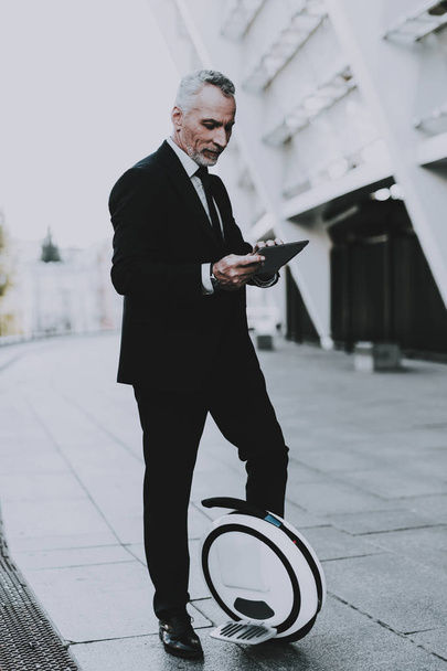 Businessman is Using a Tablet PC. Businessman is Old Smiling Man. Businessman is Typing on a Tablet. Man Wearing in Black Suit. Man is Standing on Monowheel near Skyscraper. Sunny Daytime. - Foto, Bild