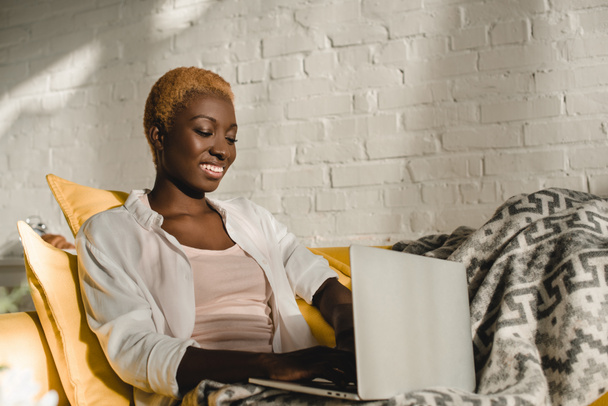 mulher americana africana alegre usando laptop no sofá amarelo
  - Foto, Imagem