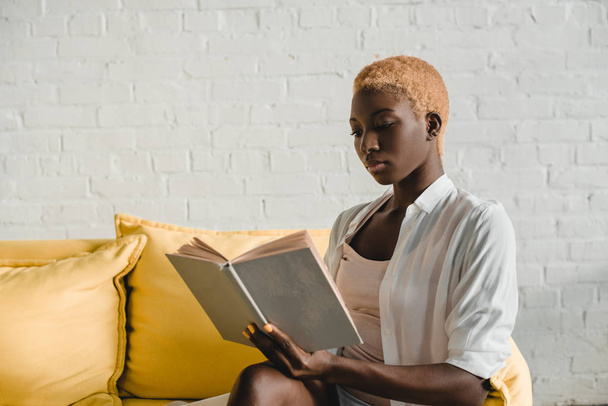 attractive african american woman with short hair reading book on yellow sofa  - Zdjęcie, obraz