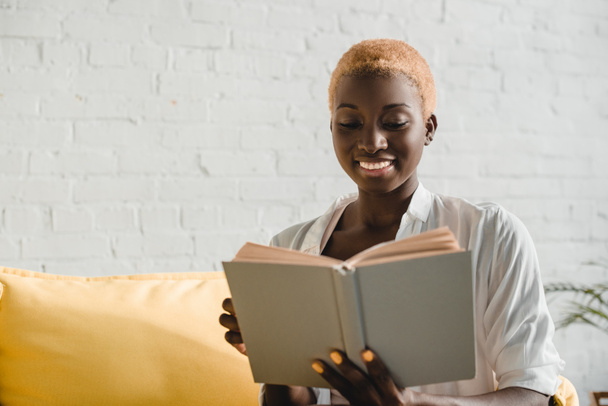fröhliche afrikanisch-amerikanische Frau mit kurzen Haaren liest Buch auf gelbem Sofa  - Foto, Bild