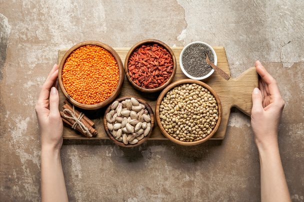 cropped view of woman holding wooden board with legumes and goji berries on rustic background - Foto, afbeelding