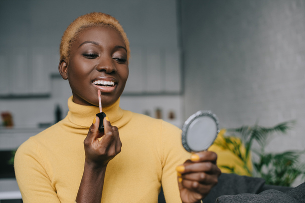cheerful african american woman applying lip gloss and looking in mirror - Fotó, kép