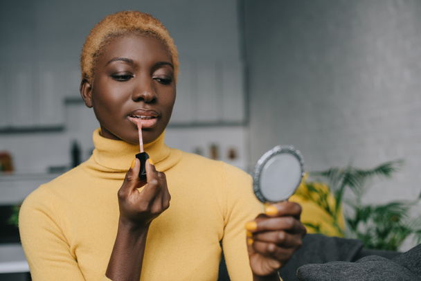 concentrated african american woman applying lip gloss and looking in mirror - Fotografie, Obrázek