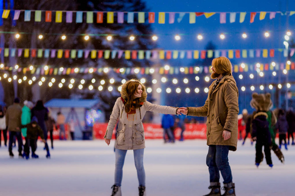 Young couple in love Caucasian man with blond hair with long hair and beard and beautiful woman have fun, active date ice skating on the ice arena in the evening city square in winter on Christmas Eve - Photo, Image
