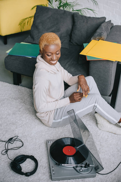 african american woman smiling and sitting on carpet near record player - Foto, afbeelding