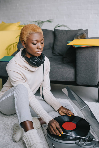 dreamy african american woman sitting on carpet in headphones near record player - Foto, afbeelding