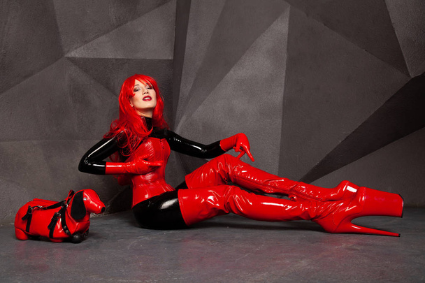 sexy mujer rizada con el pelo rojo posando en traje de goma de látex negro y fetiche corsé de pvc rojo con striptease estilo plataforma muslo botas altas y bolso de perro freak con estilo
 - Foto, Imagen
