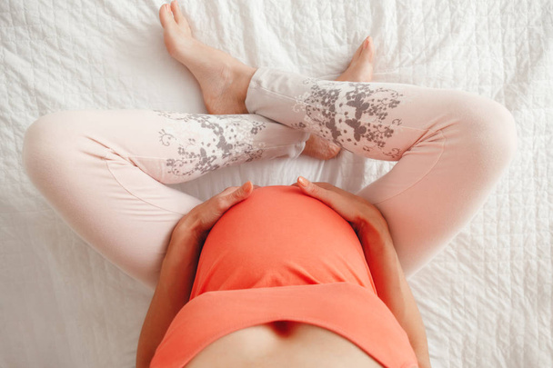 Belly of white Caucasian pregnant woman. View from top above. Expecting female lady sitting on bed in bedroom holding touching her tummy, her legs crossed. Overhead image shot, unusual angle. - Фото, изображение