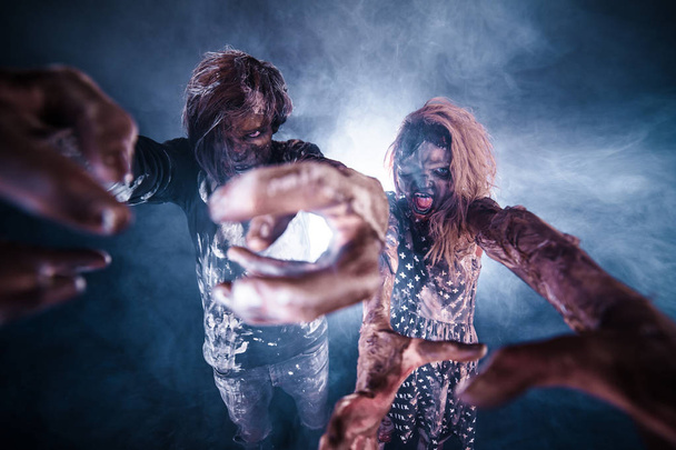 Scary zombie man and woman posing against foggy dark background with backlight - 写真・画像