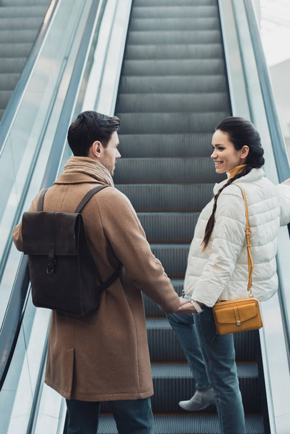 smiling couple in warm clothing holding hands and going up on escalator - Fotoğraf, Görsel