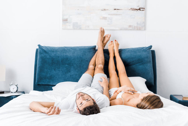 young couple lying in bed with raised legs while man looking at camera - Fotoğraf, Görsel