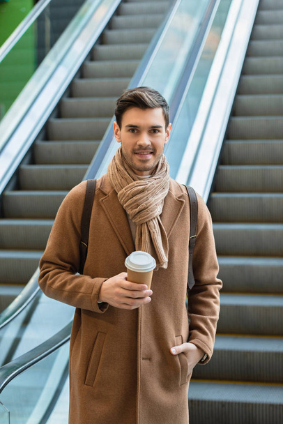 handsome man holding disposable cup and smiling on escalator - Foto, afbeelding