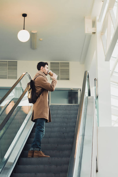 handsome man going up on escalator and drinking from disposable cup  - Photo, Image