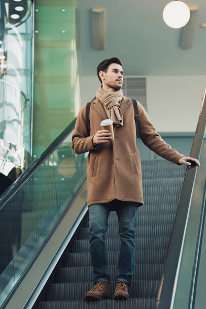 handsome man going down on escalator and holding disposable cup  - Photo, Image