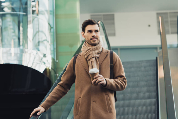 handsome man in warm clothing holding paper cup and going down on escalator - Foto, afbeelding