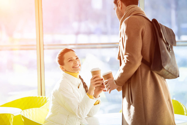 привлекательная сидящая девушка в теплой одежде и стоящий мужчина с одноразовыми чашками в кафе
 - Фото, изображение
