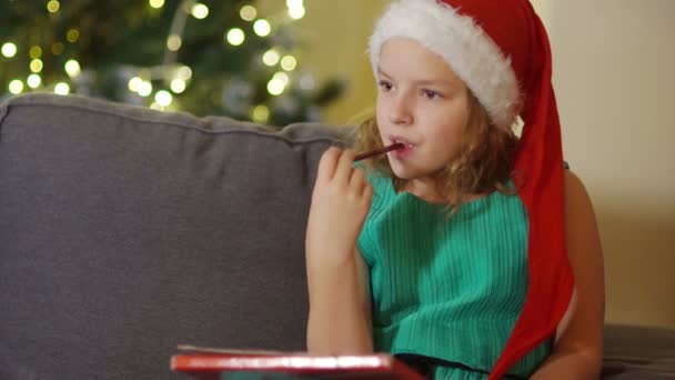A schoolgirl in an elegant green dress and Santa hat is sitting near the Christmas tree and is holding a pencil. Teen girl writes a letter to Santa Claus - Filmmaterial, Video