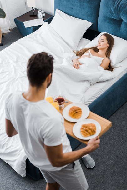 ogólny widok człowieka posiadającego drewniana Taca ze śniadaniem przy kobiecie spania w łóżku - Zdjęcie, obraz