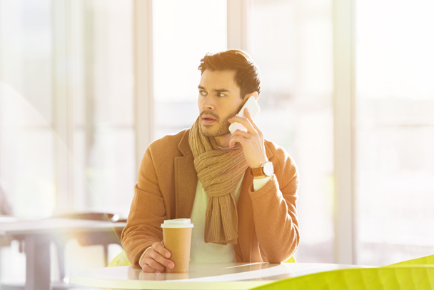 заинтересованный человек сидит за столом, держа бумажную чашку и разговаривая по смартфону в кафе
 - Фото, изображение