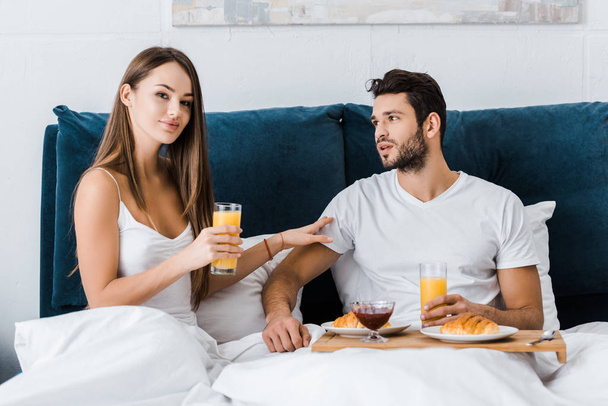 young woman holding glass of orange juice and touching boyfriend in bed - Photo, Image
