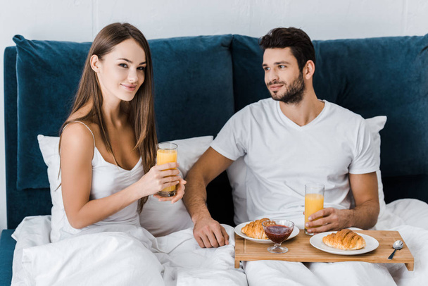 young woman holding glass of orange juice while boyfriend sitting with wooden tray - Photo, Image