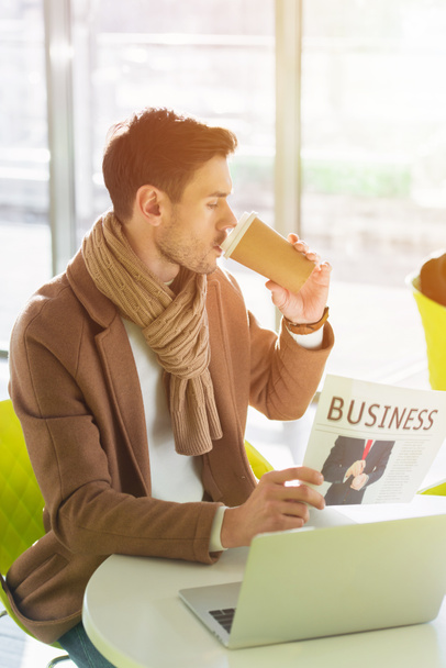 handsome businessman sitting at table, drinking coffee from paper cup and reading business newspaper in cafe - Photo, Image