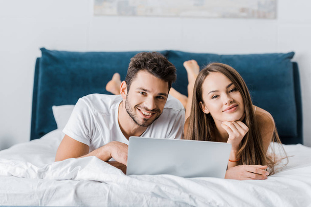 young smiling couple lying in bed with laptop and looking at camera  - Foto, imagen