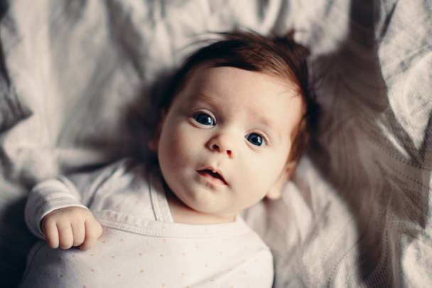 Portret zbliżenie ładny ładny zabawny biały Kaukaska brunetka małe dziecko noworodków z niebieskie oczy szare, leżąc na łóżku patrząc w aparacie. Autentyczny styl życia szczery prawdziwe życie. Stonowanych z filmu filtry. - Zdjęcie, obraz