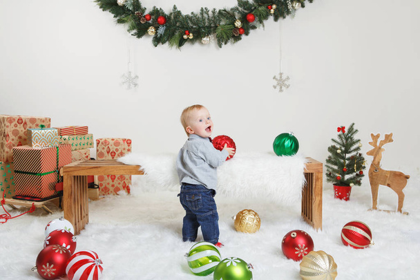 Portrait of Caucasian child baby in blue grey shirt and jeans standing on white fluffy rug celebrating Christmas or New Year. Little cute toddler boy in studio with holiday decoration ornaments. - Foto, Imagem