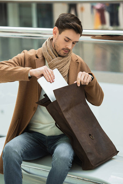 handsome man sitting on bench and putting laptop in bag - Foto, Imagen