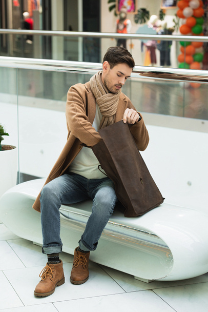 handsome man sitting on bench and holding bag in shopping mall - 写真・画像