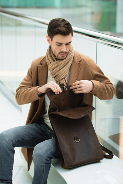 handsome man sitting on bench and opening bag in shopping mall - Fotó, kép