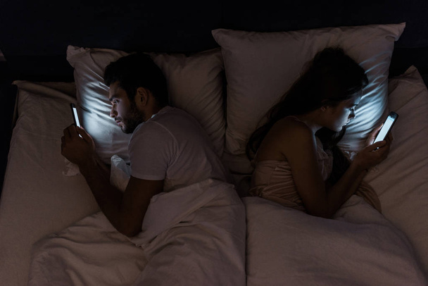 вид сверху на молодую разочарованную пару, лежащую в постели ночью и использующую смартфоны
 - Фото, изображение