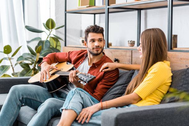young handsome man sitting on sofa in living room and playing acoustic guitar for girlfriend - Photo, Image
