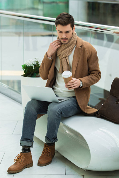 handsome man with laptop sitting on bench and holding disposable cup in shopping mall - Fotó, kép
