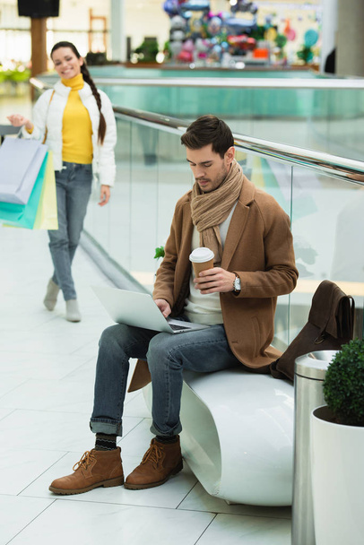 szép ember ül a padon, betöltő eldobható pohár és laptop billentyűzet gépelés bevásárlóközpontban - Fotó, kép